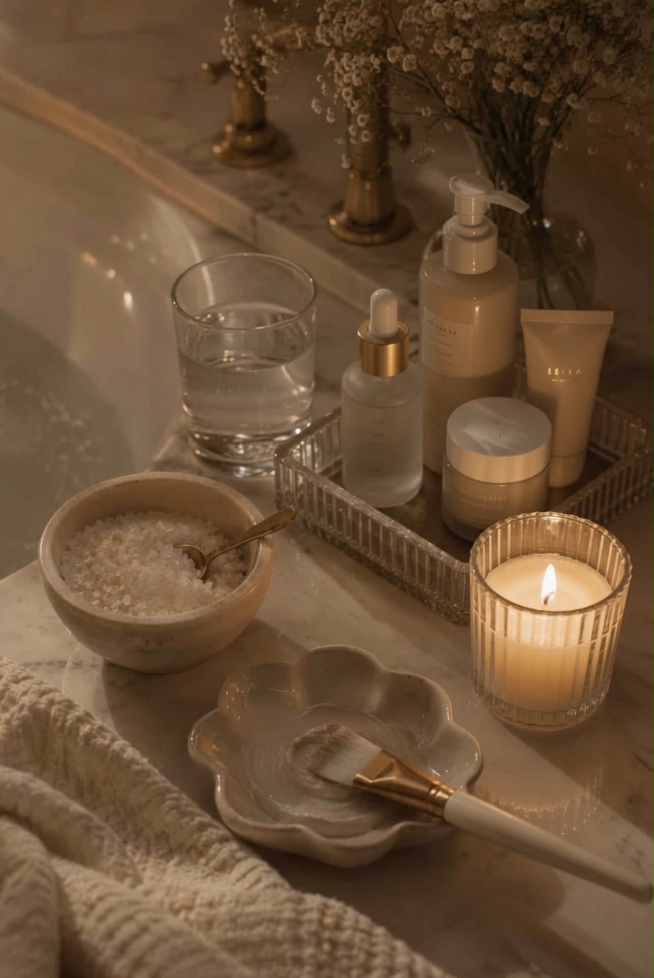 Candlelit bath tray with Epsom salts, skincare oils, a face mask brush and a lit candle beside a warm bath